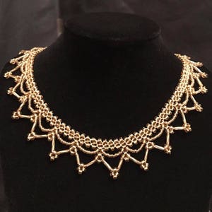 May include: A gold-toned necklace with an intricate, openwork pattern. The necklace has a series of connected, beaded components creating a scalloped edge. It is displayed on a black mannequin.