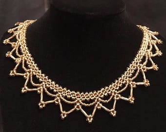 May include: A gold-colored necklace with a delicate, openwork design. The necklace features a series of connected, beaded elements forming a scalloped edge. The necklace is displayed on a black mannequin.