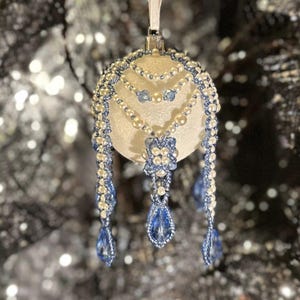 May include: A cream Christmas bauble with detailed beadwork. It features strands of white and blue beads, a floral design, and teardrop dangles. A white ribbon is attached for hanging. The bauble is a festive decoration.