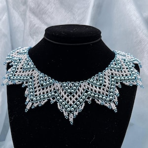 Op de afbeelding: Een opvallende ketting van witte en blauwgroene kralen. Het ingewikkelde ontwerp toont een geometrisch patroon met een geschulpte rand. De ketting wordt getoond op een zwarte mannequin buste tegen een lichtblauwe achtergrond.