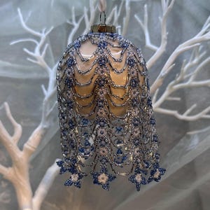 Può includere: Un ornamento a forma di campana con una base liscia color crema, ricoperta da una complessa rete intrecciata a mano di piccole perline di vetro argentate, bianche traslucide e blu scuro, rifinita con un tappo metallico e un gancio.