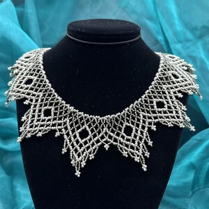 Op de afbeelding: Een zilveren kralenketting met een geometrisch ontwerp wordt getoond op een zwarte mannequin. De ketting heeft een ruitvormig patroon met een geschulpte rand. De kralen zijn klein en gelijkmatig verdeeld, wat een delicate en ingewikkelde uitstraling creëert.