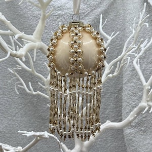 May include: A spherical cream-colored ornament featuring vertical bands of woven white pearls and gold beads. The base of the ornament is adorned with long, dangling fringe strands composed of alternating white pearls and small gold metallic beads.
