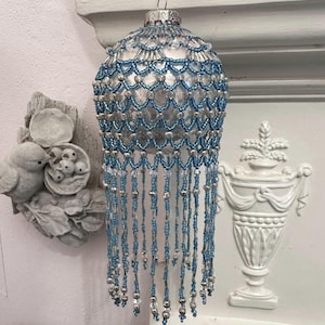 May include: A silver Christmas ornament with blue beaded detailing and dangling fringe. The ornament has a silver cap and is hanging from a silver hook. The beaded design is in a scalloped pattern. The fringe is made of silver and blue beads.