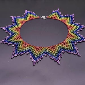 Puede incluir: Un collar de cuentas con un diseño de arcoíris. El collar tiene forma de estrella y un cierre plateado. Los colores van del rojo al amarillo, verde, azul y morado.