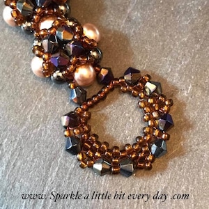 Pode incluir: Close-up de um bracelete de contas com contas marrons e roxas iridescentes. O bracelete é feito com pequenas contas de sementes e contas facetadas maiores. As contas são dispostas em um padrão circular, criando um design único e intrincado.