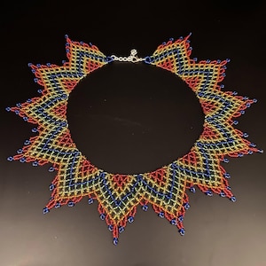 Op de afbeelding: Een kralenketting met een zonnesterontwerp. De ketting heeft een herhalend patroon van rode, gele en blauwe kralen. De ketting heeft een zilveren sluiting en wordt gepresenteerd op een zwart oppervlak.