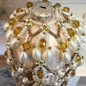 May include: A decorative Christmas bauble with a cream-coloured base, adorned with white, gold, and amber-coloured beads and faux pearls. The bauble has a silver metal cap for hanging. The design features beaded dangles and floral accents.