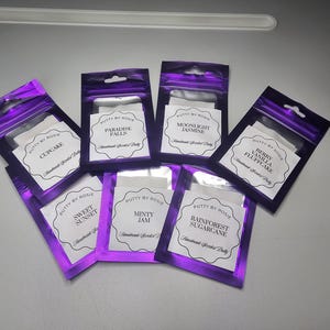 May include: Several purple foil pouches filled with scented putty. Each pouch features a white label with the text "PUTTY BY ROSIE" and a scent name such as "Cupcake" or "Paradise Falls". The pouches are displayed on a light grey surface.
