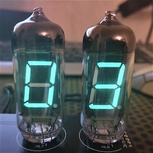 Może przedstawiać: Dwa przezroczyste szklane lampy Nixie z zielonymi świecącymi cyframi, wyświetlające cyfry 0 i 1. Lampy są oznaczone "HL71" i "HLB1".