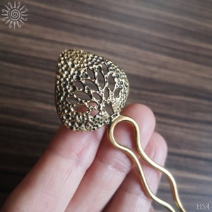 Op de afbeelding: Een goudkleurige metalen haarkam met een decoratief bovendeel met een gehamerd, getextureerd oppervlak en een uitgesneden boomontwerp. Het haaraccessoire heeft een tweekoppige, golvende steel die uit de basis van het ornament voortkomt.