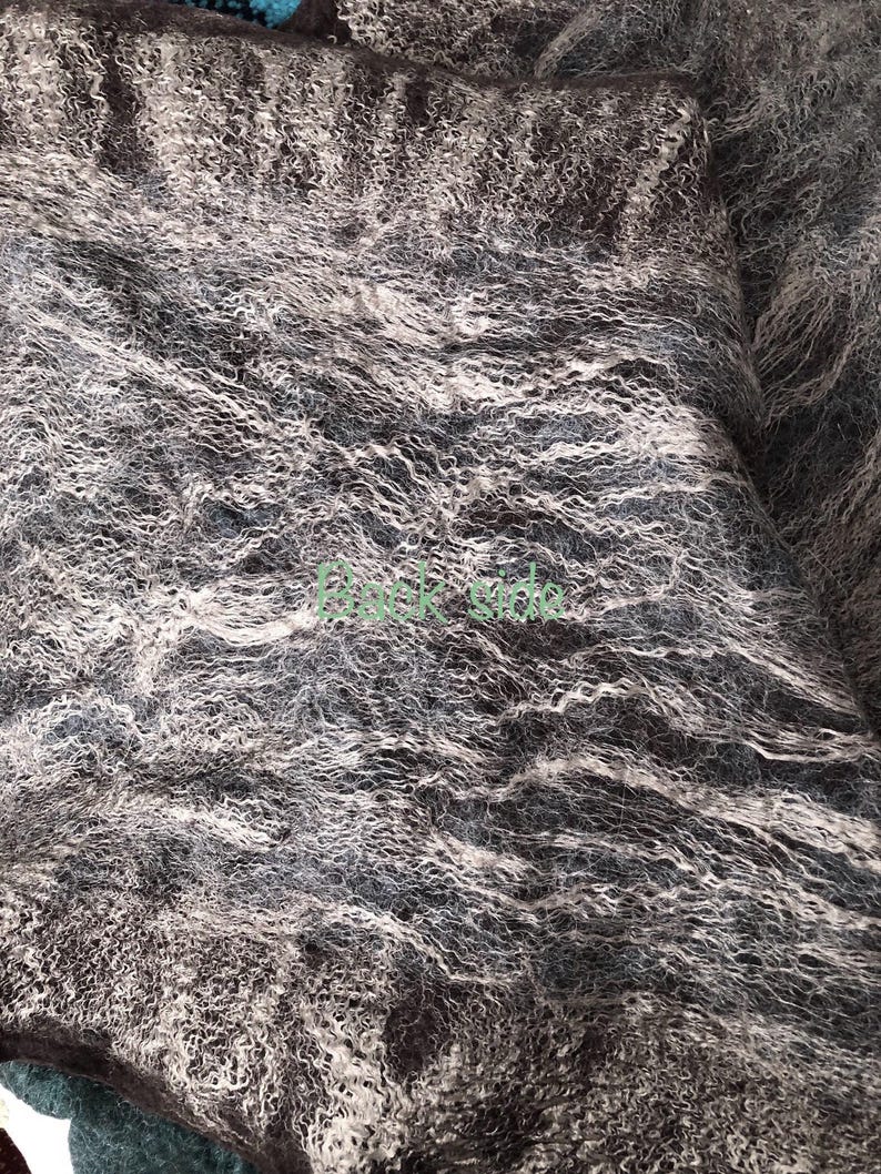 May include: Close-up of a textured fabric in shades of gray, brown, and white. The material has a wavy, almost fur-like appearance, with a mix of light and dark tones creating depth and visual interest. The fabric appears soft and luxurious.
