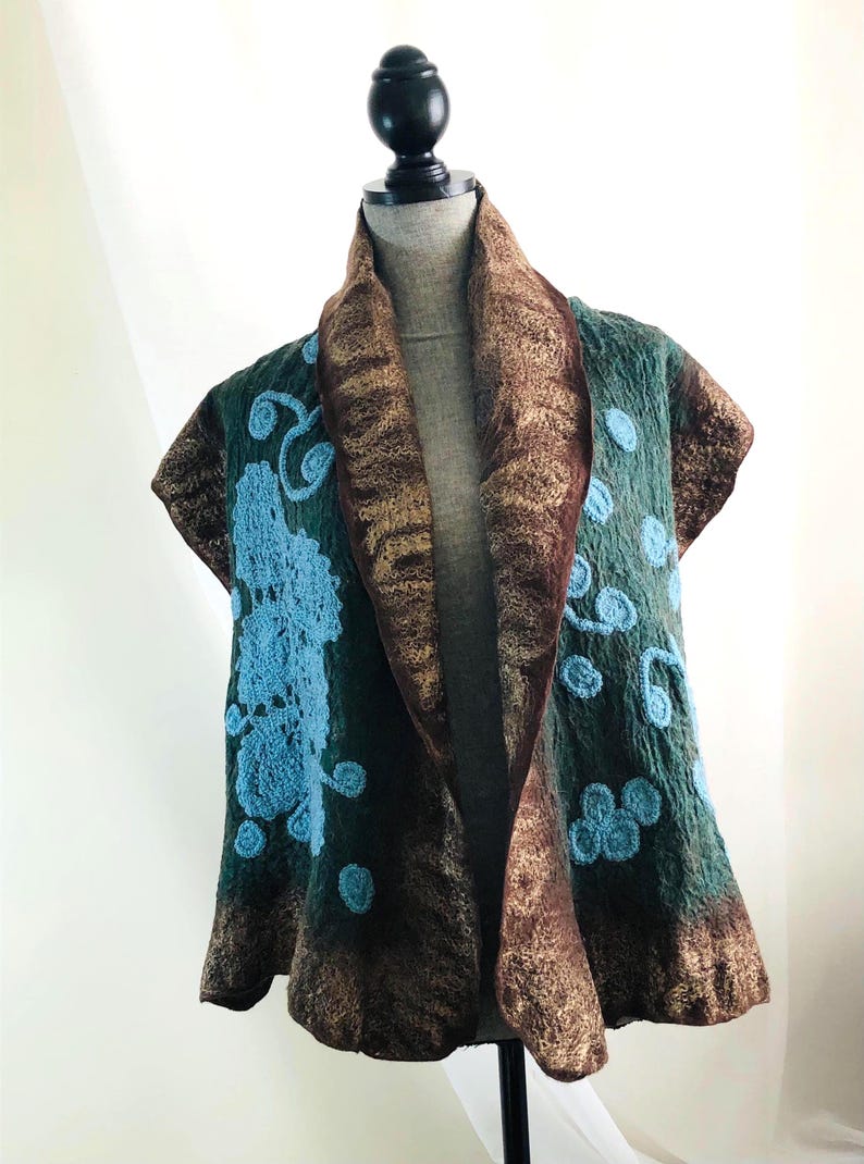 May include: A teal and brown felted vest with a shawl collar. The vest features light blue floral embroidery and a brown trim. The vest is displayed on a mannequin.