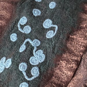May include: Close-up of a textile with a textured, felted appearance. The fabric is primarily dark gray and brown, with light blue crocheted floral embellishments. The design features various sizes of circular and curved shapes.