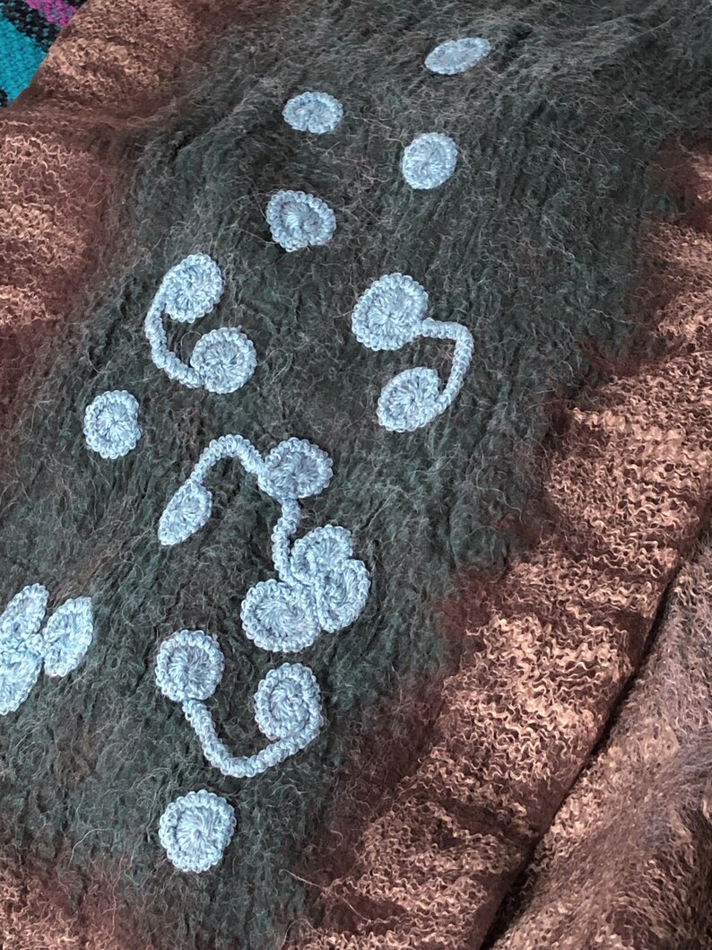 May include: Close-up of a textile with a textured, felted appearance. The fabric is primarily dark gray and brown, with light blue crocheted floral embellishments. The design features various sizes of circular and curved shapes.