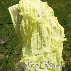 May include: A bright yellow, crinkled, sheer fabric shawl draped over a wooden fence post. The fabric is very light and airy, and it has a delicate, almost translucent quality. The text "Galina Shawls" is visible in the lower right corner of the image.