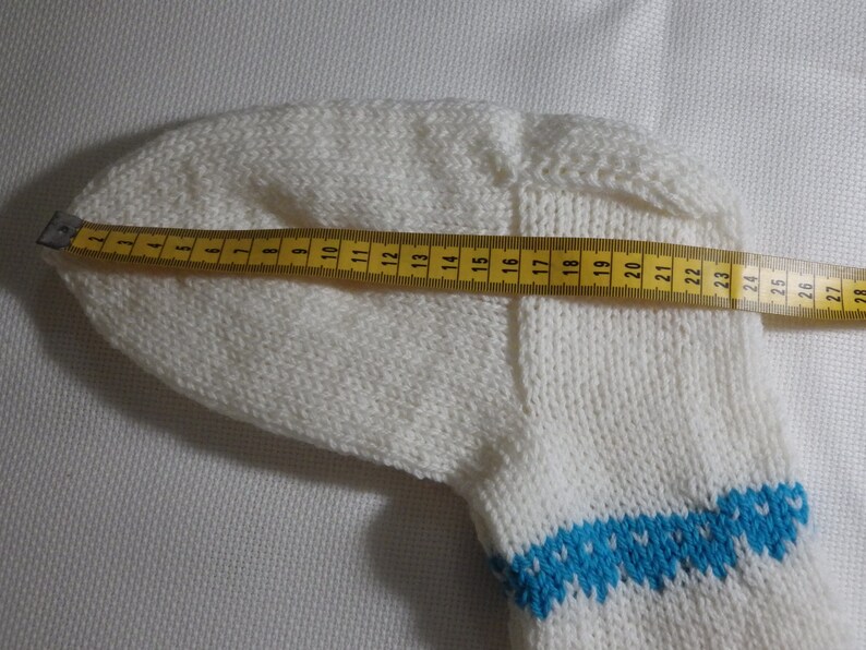 K&ouml;nnte beinhalten: Wei&szlig;e Stricksocke mit einem blauen und wei&szlig;en gemusterten B&uuml;ndchen. Die Socke wird mit einem gelben Ma&szlig;band gemessen, das eine L&auml;nge von 71 cm zeigt.
