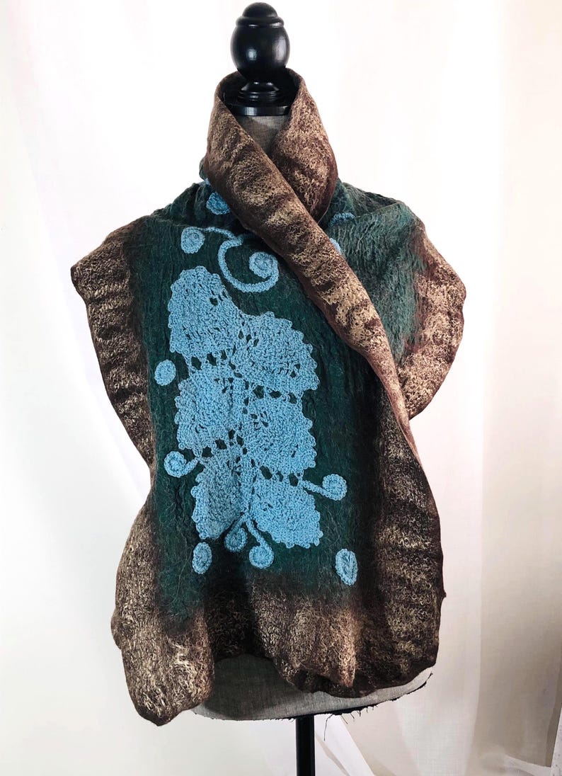 May include: A handcrafted felted wool scarf in shades of green and brown, adorned with a light blue crocheted floral design. The scarf is draped over a mannequin, showcasing its unique texture and artistic detail.
