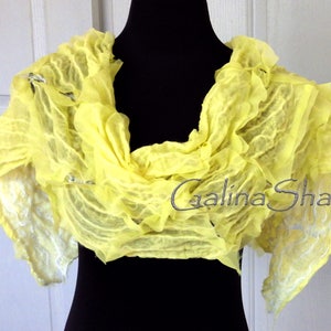 May include: A bright yellow silk scarf with a ruffled texture. The scarf is draped over a black mannequin and has a white, textured design on the edges. The text "Galina Shawls" is visible on the scarf.