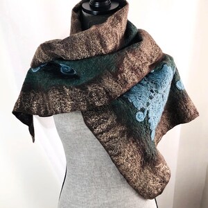 May include: A felted wool scarf in shades of brown, green, and blue. The scarf features a textured design with a blue floral pattern on one corner. The scarf is draped over a mannequin.