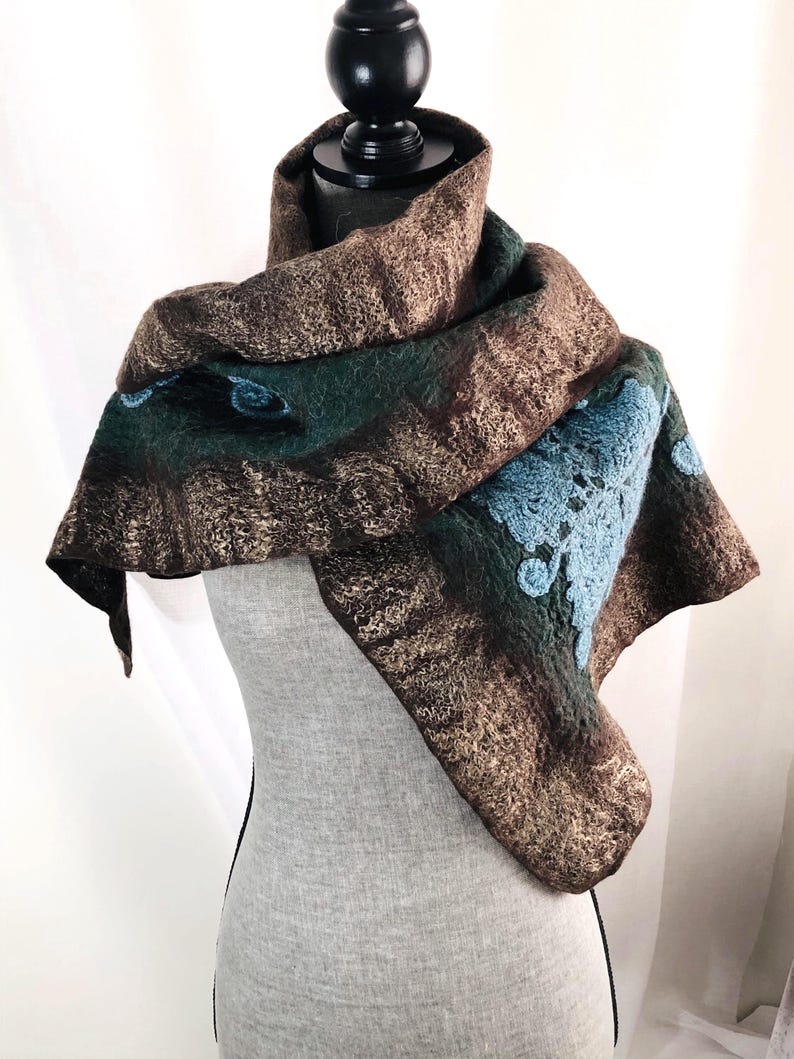May include: A felted wool scarf in shades of brown, green, and blue. The scarf features a textured design with a blue floral pattern on one corner. The scarf is draped over a mannequin.