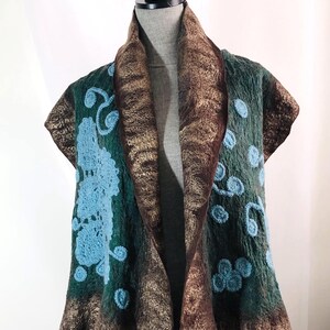 May include: A green and brown felted vest with blue floral embroidery. The vest has a shawl collar and is displayed on a mannequin. The edges are trimmed with a metallic gold fabric.