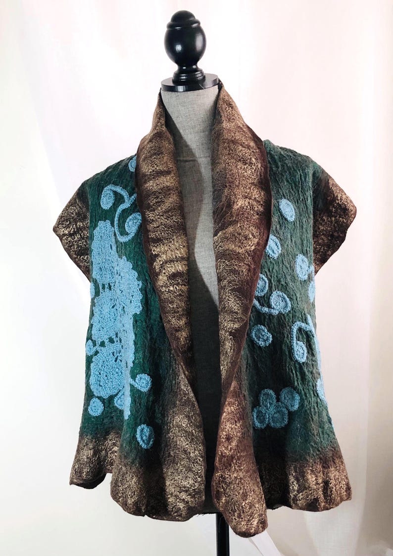 May include: A green and brown felted vest with blue floral embroidery. The vest has a shawl collar and is displayed on a mannequin. The edges are trimmed with a metallic gold fabric.
