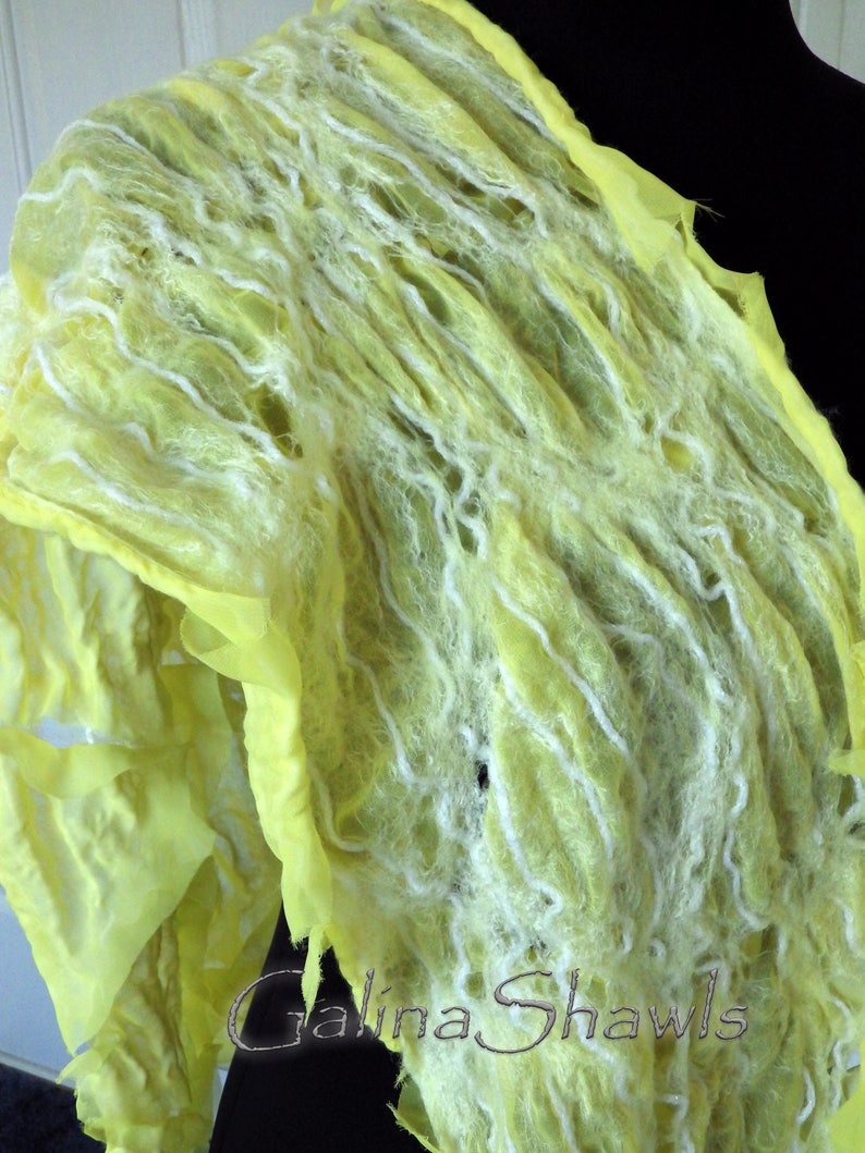 May include: A yellow and white wool shawl with a textured, woven design. The shawl is draped over a black surface. The text "Galina Shawls" is visible in the lower right corner of the image.
