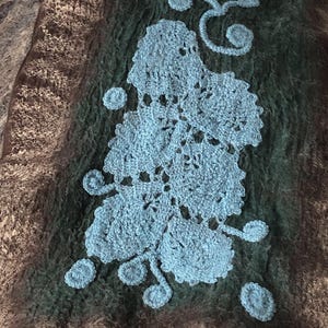 May include: Textured textile in shades of brown, green, and blue. A large, light blue crocheted floral design is centered on the dark green section. The textile has a soft, fuzzy appearance.