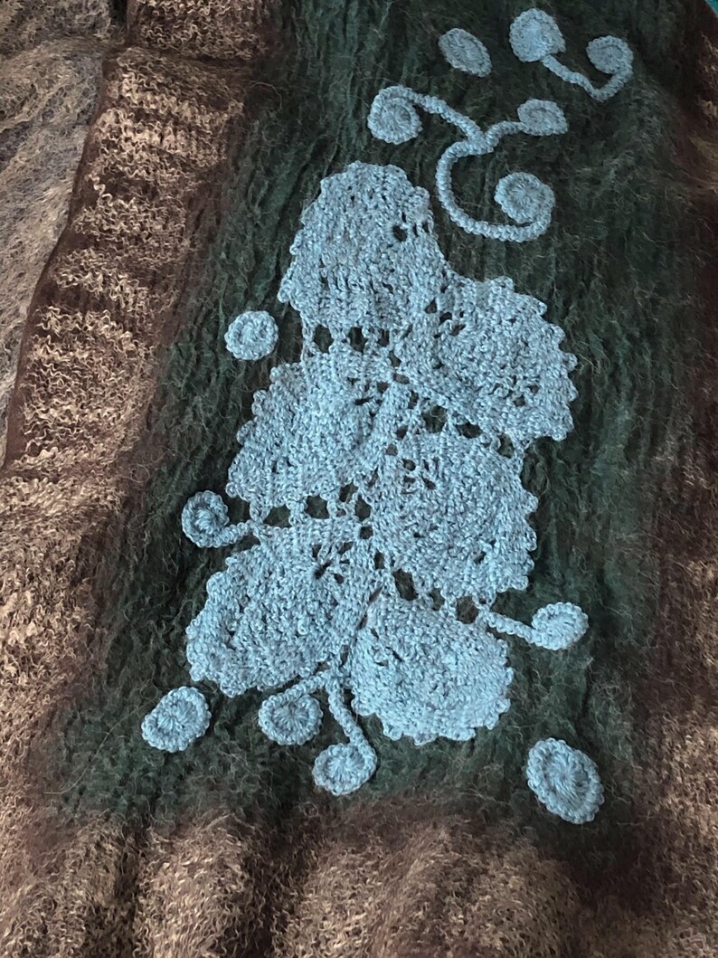 May include: Textured textile in shades of brown, green, and blue. A large, light blue crocheted floral design is centered on the dark green section. The textile has a soft, fuzzy appearance.