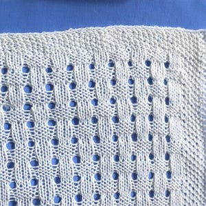 May include: Close-up of a white knitted textile with a repeating pattern of woven sections and small, evenly spaced holes. The texture is visible, and the background is a solid blue color. The item appears to be a blanket or a piece of fabric.