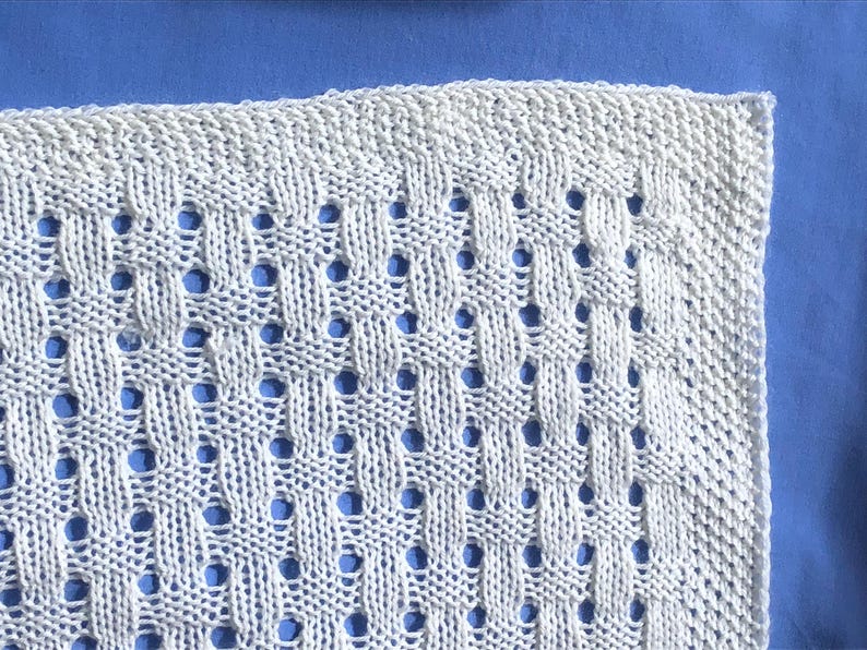 May include: Close-up of a white knitted textile with a repeating pattern of woven sections and small, evenly spaced holes. The texture is visible, and the background is a solid blue color. The item appears to be a blanket or a piece of fabric.