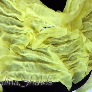 May include: A yellow sheer fabric shawl with a crinkled texture. The fabric is draped and folded, creating a soft and flowing look. The text "Galina Shawls" is visible at the bottom of the image.