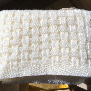 May include: Cream-colored knitted blanket with a woven basket weave pattern. The blanket is rectangular and appears to be made of soft yarn. The texture is visible, with a repeating pattern of raised and recessed areas.