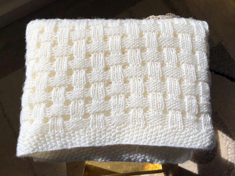 May include: Cream-colored knitted blanket with a woven basket weave pattern. The blanket is rectangular and appears to be made of soft yarn. The texture is visible, with a repeating pattern of raised and recessed areas.