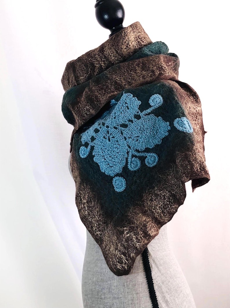 May include: A brown and teal felted scarf with a blue crocheted floral design. The scarf is draped over a mannequin head, showcasing its texture and shape. The scarf's design features a unique blend of colors and textures.