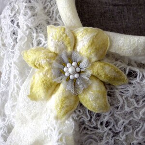 以下が含まれることがあります： 白いクロシェのショールに、黄色の花のブローチが付いている。花の中心は白く、真珠の装飾が施されている。