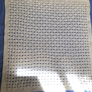 May include: A beige knitted blanket with a woven pattern. The blanket is rectangular and has a textured surface. The blanket is made of a soft material and is suitable for use as a baby blanket or throw.