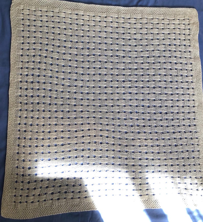 May include: A beige knitted blanket with a woven pattern. The blanket is rectangular and has a textured surface. The blanket is made of a soft material and is suitable for use as a baby blanket or throw.