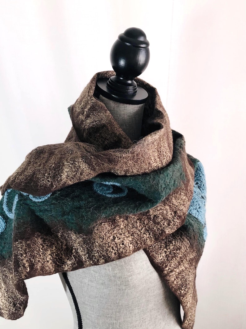 May include: A brown, green, and blue felted wool scarf draped over a mannequin. The scarf features a textured, patterned design with shades of brown, green, and blue. The scarf is made of wool.