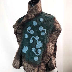 May include: A felted shawl in shades of teal and brown, adorned with light blue embroidered designs. The shawl has a textured appearance and is displayed on a mannequin. The edges are trimmed with a metallic gold fabric.