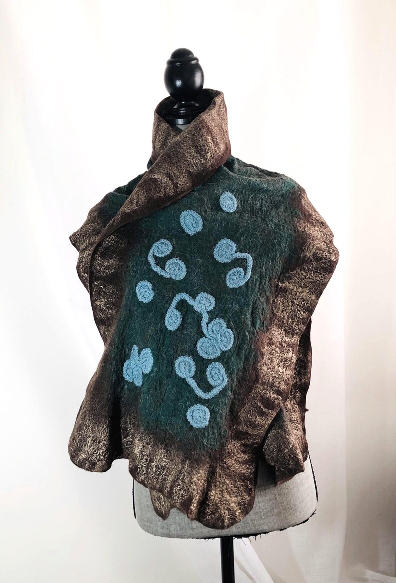 May include: A felted shawl in shades of teal and brown, adorned with light blue embroidered designs. The shawl has a textured appearance and is displayed on a mannequin. The edges are trimmed with a metallic gold fabric.