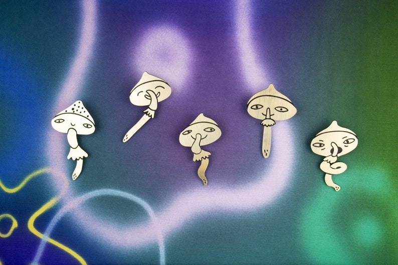 May include: Five wooden mushroom-shaped ornaments with whimsical faces. Each mushroom has a different expression, including a smiling face, a face with its finger to its lips, and a face with its finger to its nose. The ornaments are painted in a light brown colour and have black details.