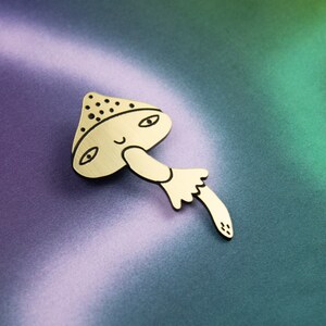 May include: A gold-coloured wooden mushroom pin with a smiling face and black eyes. The mushroom has a brown and white spotted cap and a stem with a white ruffle.