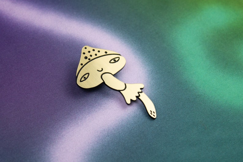 May include: A gold-coloured wooden mushroom pin with a smiling face and black eyes. The mushroom has a brown and white spotted cap and a stem with a white ruffle.
