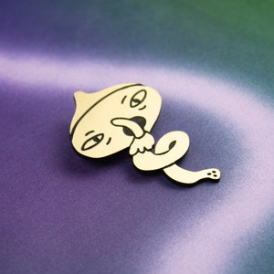 May include: A wooden pin featuring a cartoon character design. The character has a round head with a wide mouth and a tongue sticking out. The character is wearing a hat and has a long, thin body that is curled up.