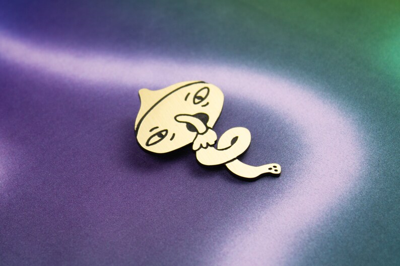 May include: A wooden pin featuring a cartoon character design. The character has a round head with a wide mouth and a tongue sticking out. The character is wearing a hat and has a long, thin body that is curled up.