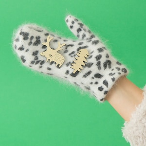 May include: A white and black spotted mitten with wooden cutouts of a reindeer and a Christmas tree. The mitten is on a hand with a green background. The mitten is made of a soft, fuzzy material.