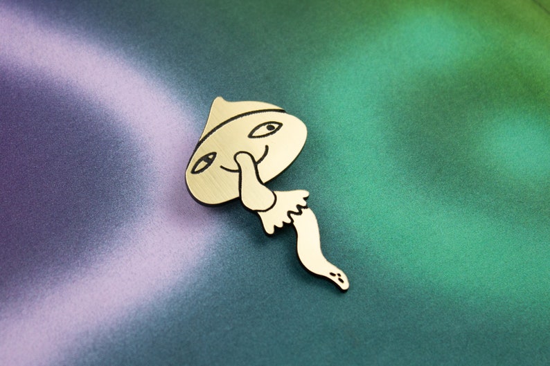May include: A gold-coloured metal pin shaped like a mushroom with a smiling face and a long, thin stem. The mushroom is wearing a hat.