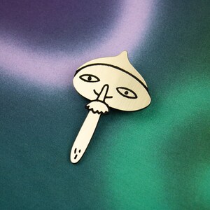 May include: A gold-coloured metal pin shaped like a mushroom with a face. The mushroom has a black outline and black eyes, a nose, and a moustache.